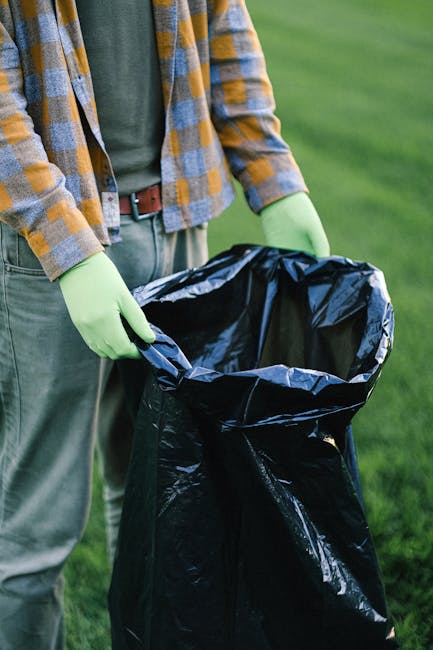 The image shows a person standing outdoors on a grassy area, holding an open black trash bag with both hands, which are protected by bright green disposable gloves. The individual is dressed in a casual outfit consisting of a grey t-shirt and a plaid shirt with shades of yellow, blue, and grey, along with light grey trousers. The black plastic bag appears to be large and flexible, suitable for collecting waste or rubbish. The background features lush green lawn, suggesting a garden or park environment, and the lighting indicates daytime with natural light highlighting the scene. This context relates to rubbish removal or waste disposal activities, possibly involving independent collection or on-site clearance managed by a waste management service like Rubbish Removal Dulwich, operating within the category of rubbish disposal solutions.