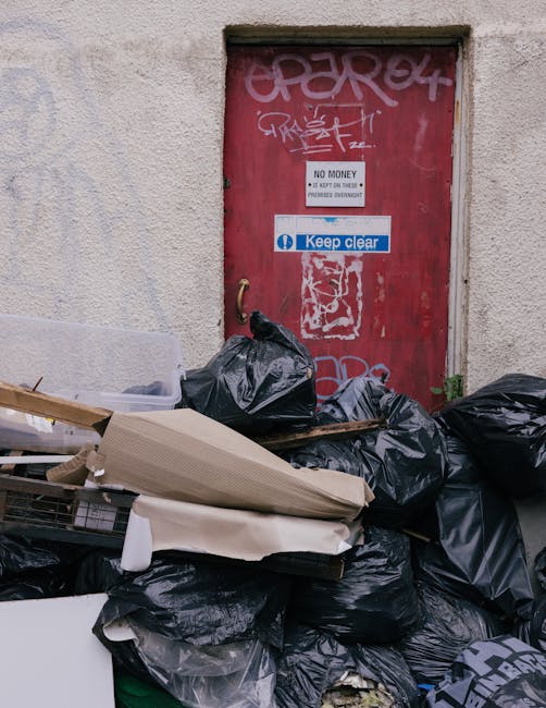 A large pile of black plastic rubbish bags and discarded cardboard and packaging materials are stacked in front of a red metal door mounted on a rough-textured beige wall. The bags appear full and tightly sealed, some are leaning against each other, with a few resting on top of cardboard and wooden pieces. The door is weathered, with graffiti tags and stickers attached, including one that reads 'Keep clear' in blue and white and another warning sign indicating 'No money, keep door closed.' The surrounding environment suggests an outdoor urban area, possibly a service or back entrance, with natural daylight illuminating the scene. This image visually represents the concept of private waste collection or rubbish removal, similar to services offered by companies like Rubbish Removal Dulwich, which provide alternative waste handling solutions beyond municipal disposal, emphasizing the importance of proper rubbish management in residential or commercial settings.