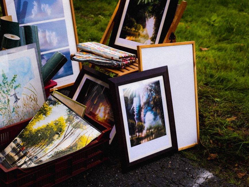 A collection of framed landscape photographs and artworks arranged outdoors on a patch of grass, with some leaning against each other and others placed on a red plastic crate. The images feature scenes of trees, water, and nature, with a range of colours including greens, blues, and golden sunset hues. Several of the frames are wooden with light or dark finishes, while others have plain white or black borders. On top of the crate, there are two neatly folded fabric cloths with colorful patterns. In the background, additional framed artworks are visible, some propped on wooden easels or partially obscured by other pieces. The scene appears to be part of an outdoor art or photography sale, with natural daylight illuminating the area, highlighting the textures and colours of the framed works, and creating a casual, temporary display typical for scheduled rubbish removal or private disposal of unwanted items, in line with services provided by Rubbish Removal Dulwich.