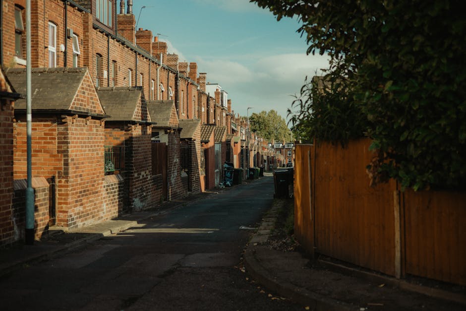 A narrow residential alleyway lined with red-brick terraced houses on the left, featuring pitched roofs with chimney stacks and small front steps, casting long shadows on the pavement. On the right, a tall wooden fence runs parallel to the street, partially obscuring a garden area with dense green foliage. Several black wheelie bins are positioned along the curb, likely for waste collection or private rubbish disposal, reflecting typical outdoor waste containers in urban environments. The pavement appears clean but slightly weathered, with a few scattered fallen leaves. In the background, the alley extends into the distance with more houses visible under a partly cloudy blue sky, illuminated by late afternoon sunlight, suggesting an environment where independent rubbish removal services could be used for waste collection, complementing or substituting council-provided rubbish collection for local households.