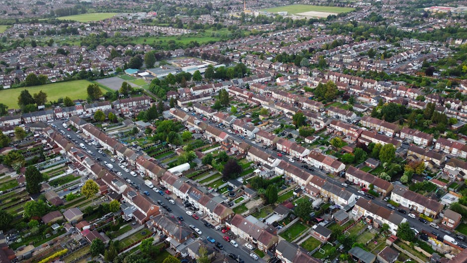 An aerial view of a residential suburban area showing rows of terraced houses with small front gardens and driveways, parked cars along the roads, and a mix of green trees and grassy patches interspersed throughout the neighbourhood. The houses have brick walls and tiled roofs, with some featuring small front porches and variously colored doors. In the background, there are larger green spaces, sports fields, and scattered trees, indicating recreational areas. The scene is illuminated by natural daylight, creating a clear and detailed perspective of the community layout, typical of urban fringe or outer suburban districts where private waste collection services such as Rubbish Removal Dulwich operate. The image captures the organized nature of a typical residential street, highlighting the density of housing while the greenery provides a contrasting natural element common in neighbourhood waste management and rubbish removal contexts.
