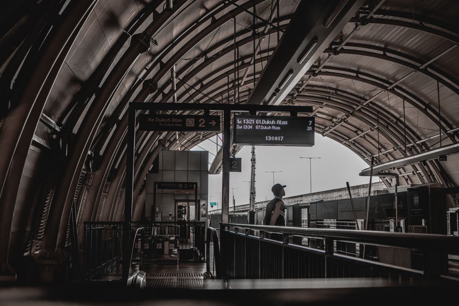 Inside an underground train station with an arched metallic ceiling structure and a platform area. A woman with a backpack is standing near the edge of the platform, facing away from the camera, looking towards the train tracks. Above her, there are electronic display boards showing train departure times and destinations, including references to Dukenh Avenue. The environment features a combination of dark metallic surfaces and natural light filtering through the station's entrance or open sections, creating contrasting shadows and highlights. Rubbish or clutter is not visible in this image, but the setting suggests a location where efficient waste management and rubbish removal services, such as those offered by Rubbish Removal Dulwich, could support maintaining clean public transportation environments by facilitating private or independent waste clearance of station areas, especially in the context of efforts to ensure tidy and efficient transit hubs.