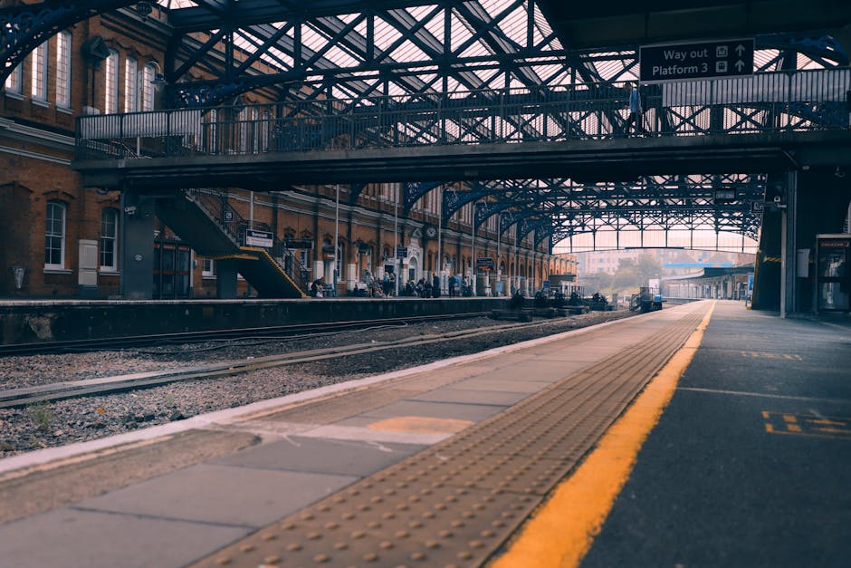 A view of East Dulwich train station platform with an arched, glass-paneled roof supported by dark metal trusses. The platform is made of concrete with a tactile yellow warning strip along the edge for indicating safe distance from the tracks. In the background, there are brick station buildings with large windows and a staircase leading to an upper level on the left side. Several passengers are visible waiting on the platform, some sitting on benches while others stand near the edge. Railway tracks run parallel to the platform, with gravel ballast visible between the rails. The station area is illuminated by natural daylight filtering through the glass roof, creating a well-lit environment that highlights the industrial architectural features. Signage overhead indicates directions for way out and platform numbers, contributing to the station's functional appearance. The scene exemplifies an active transit environment, with potential relevance for services related to station rubbish clearance or maintenance tasks by waste management companies such as Rubbish Removal Dulwich.