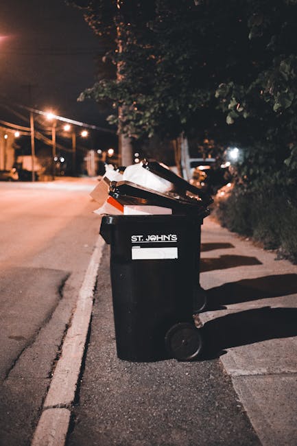 A black wheeled rubbish bin labeled 'ST. JOHN'S' is positioned on the pavement next to a curb on a quiet residential street at night. The bin's lid is open, revealing a mix of discarded cardboard boxes, paper, and plastic waste. The cardboard appears brown and slightly crumpled, with some white paper or packaging visible among the contents. The surrounding environment includes trees and dense bushes to the right, casting shadows on the sidewalk, while streetlights in the background illuminate the scene with a warm, orange glow. Several other bins or containers are visible in the distance along the street, suggesting regular refuse collection points. The scene exudes a calm, suburban atmosphere, with the bin prepared for an early morning collection as part of an alternative or private waste disposal service, such as Rubbish Removal Dulwich, catering to domestic rubbish clearance needs.