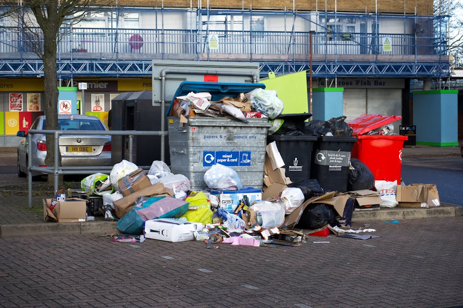 A large collection of mixed household waste, including various cardboard boxes, paper bags, plastic bags, and loose paper, is piled around multiple wheeled bins on a paved urban sidewalk. The bins, positioned in front of a row of commercial shops with signage and storefront window displays, include a grey bin labeled for mixed paper and cards, a black bin for general waste, and a bright red bin. Additional waste bags, partly black and some white, are stacked beside and on top of the bins, overflowing onto the surrounding pavement. A few small cardboard boxes and loose items are scattered on the ground nearby. Behind the waste area, a parked silver car is visible, with a metal railing separating it from the waste collection spot. In the background, a building under construction or renovation is covered in blue scaffolding, with some signage and windows partially visible. The scene is illuminated by natural daylight, highlighting the disorganized waste and contrasting it with the storefronts and urban environment, illustrating a typical scenario where independent rubbish collection may be needed for efficient clearance.