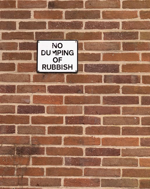 Two large black plastic rubbish bags, filled and tied at the top, are positioned on the pavement near a wooden fence with vertical slats. The bags appear to contain household waste, with some of the material bulging through their crinkled, shiny surface. To the left of the bags, a rectangular cardboard box, possibly a discarded packaging item, is lying on its side, with visible text and branding partially obscured. Behind the fence, dense foliage from trees or bushes extends upward, providing a natural backdrop. The scene is lit by natural daylight, casting soft shadows on the ground and highlighting the textures of the materials. The setting suggests an area where domestic waste has been temporarily gathered, and the presence of Rubbish Removal Dulwich indicates professional collection or disposal is part of an alternative waste handling service, consistent with non-local authority rubbish collection practices in the Dulwich area.
